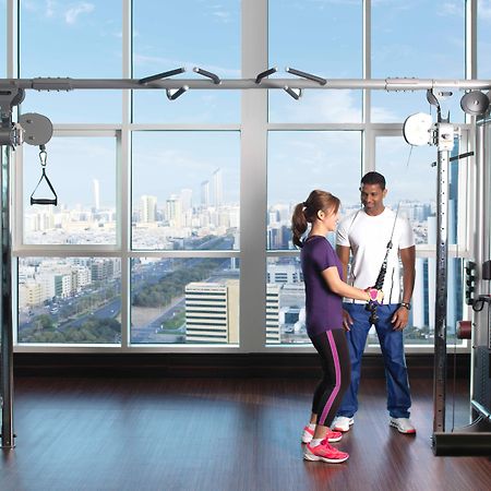 Dusit Thani Abu Dhabi Exterior photo The photo shows a modern gym setting with large windows offering a view of a cityscape. In the foreground, there are two individuals engaged in a training session. One person, a woman in athletic wear, appears to be exercising with a machine, while a