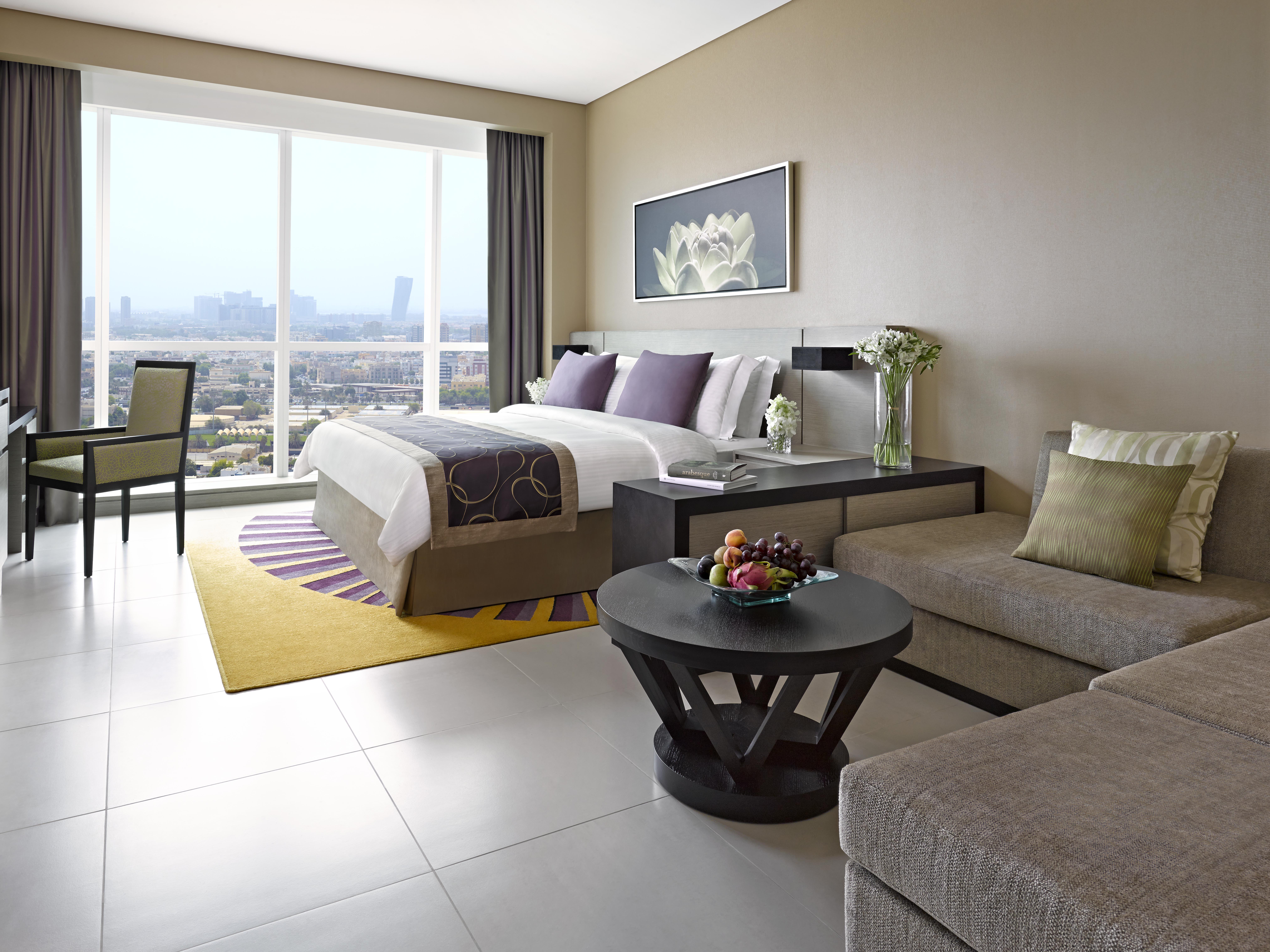 Dusit Thani Abu Dhabi Exterior photo The photo shows a modern hotel room featuring a combination of a bedroom and a living area. There is a large bed with white linens and decorative purple pillows, positioned against a wall. Next to the bed, there is a bedside table and a small vase wi