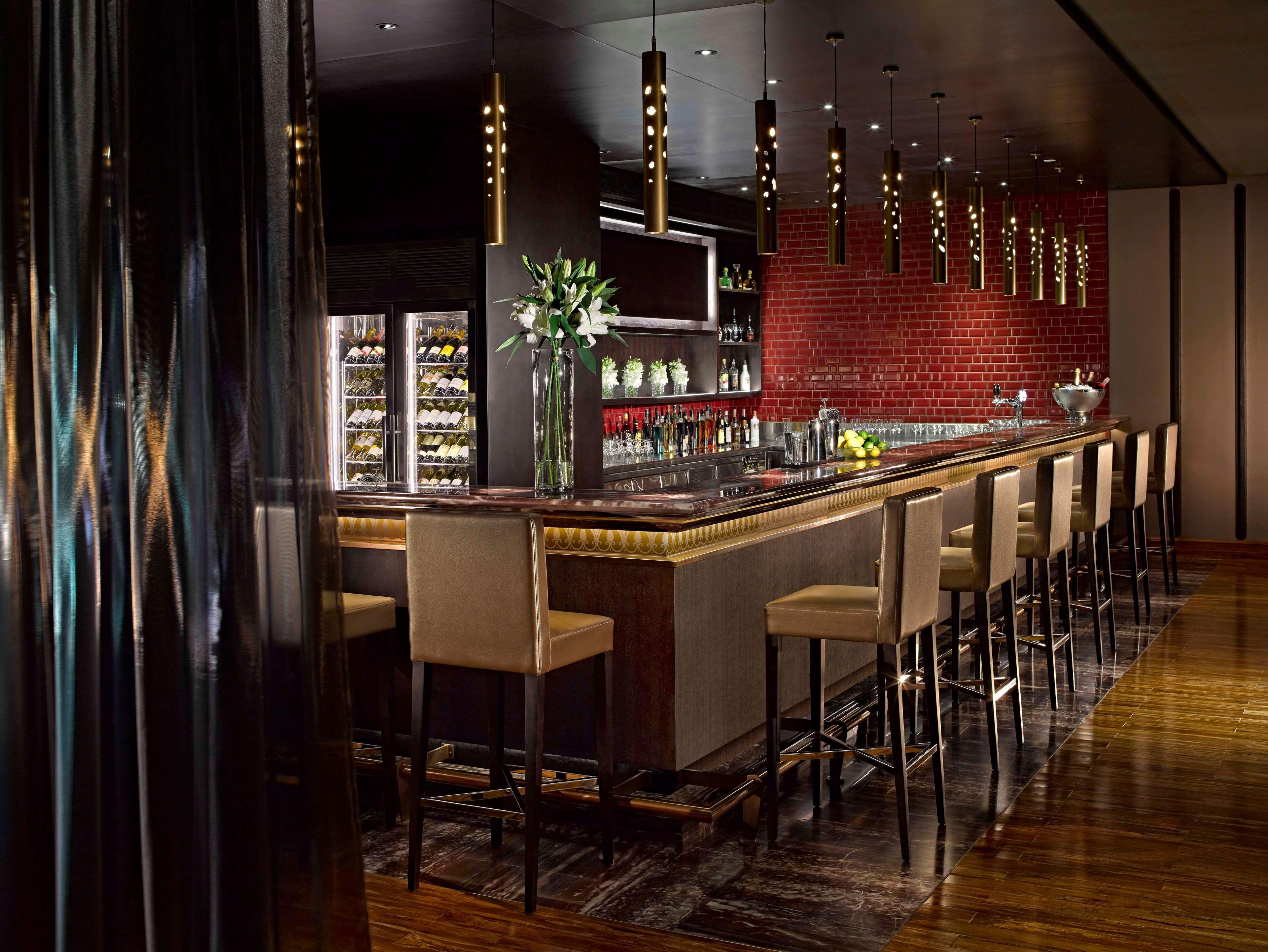 Dusit Thani Abu Dhabi Exterior photo The photo depicts a stylish and modern bar area. There is a long bar countertop with a sleek design, featuring elegant lighting fixtures hanging above it. The bar is lined with high stools, each with a light-colored upholstery. Behind the bar, there 