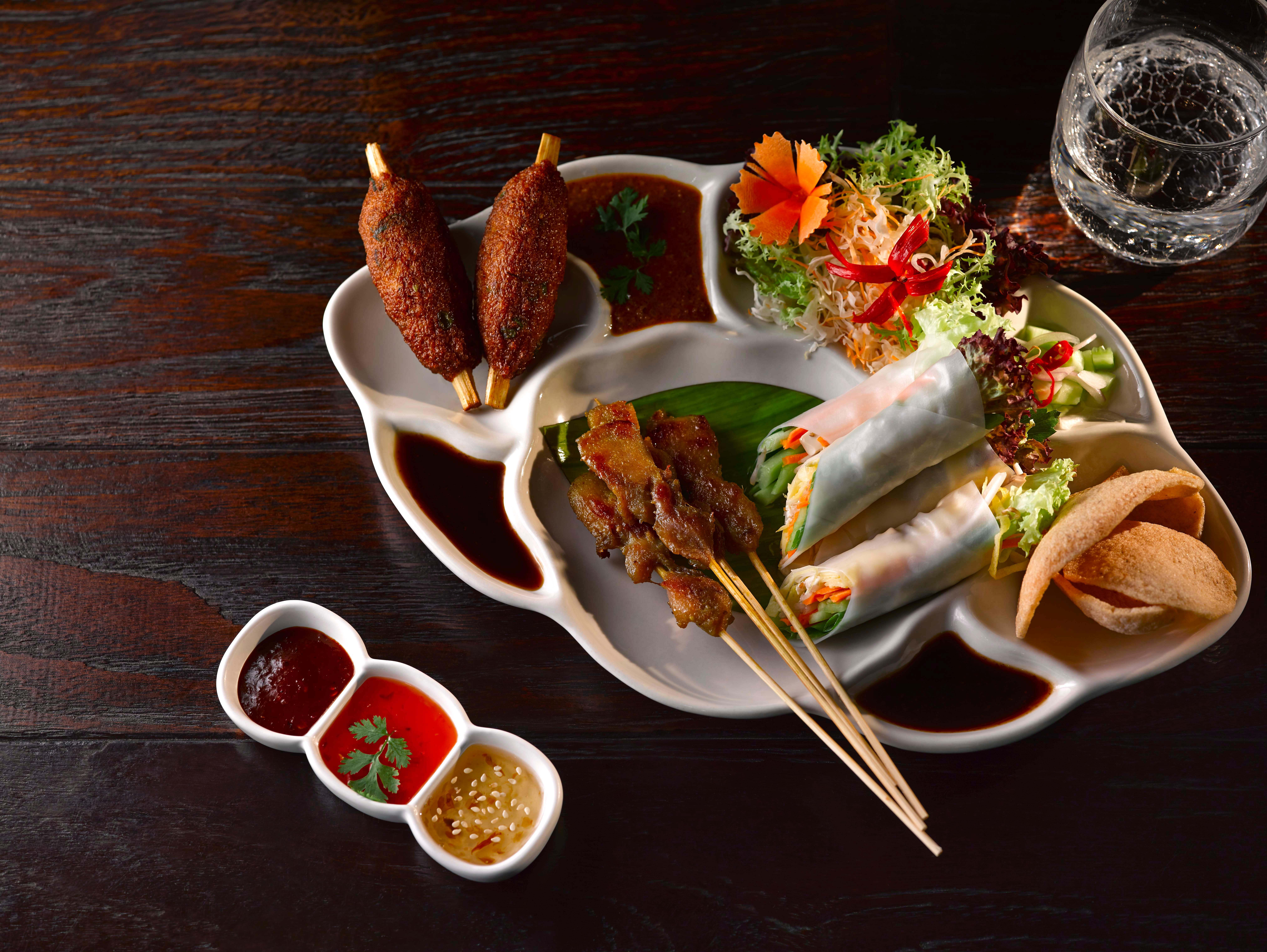 Dusit Thani Abu Dhabi Exterior photo The photo displays an elegant platter featuring various appetizers. On the left, there are two skewers, likely grilled meat. Next to them, there are fresh spring rolls filled with vegetables and herbs, accompanied by a small side of greens and some d