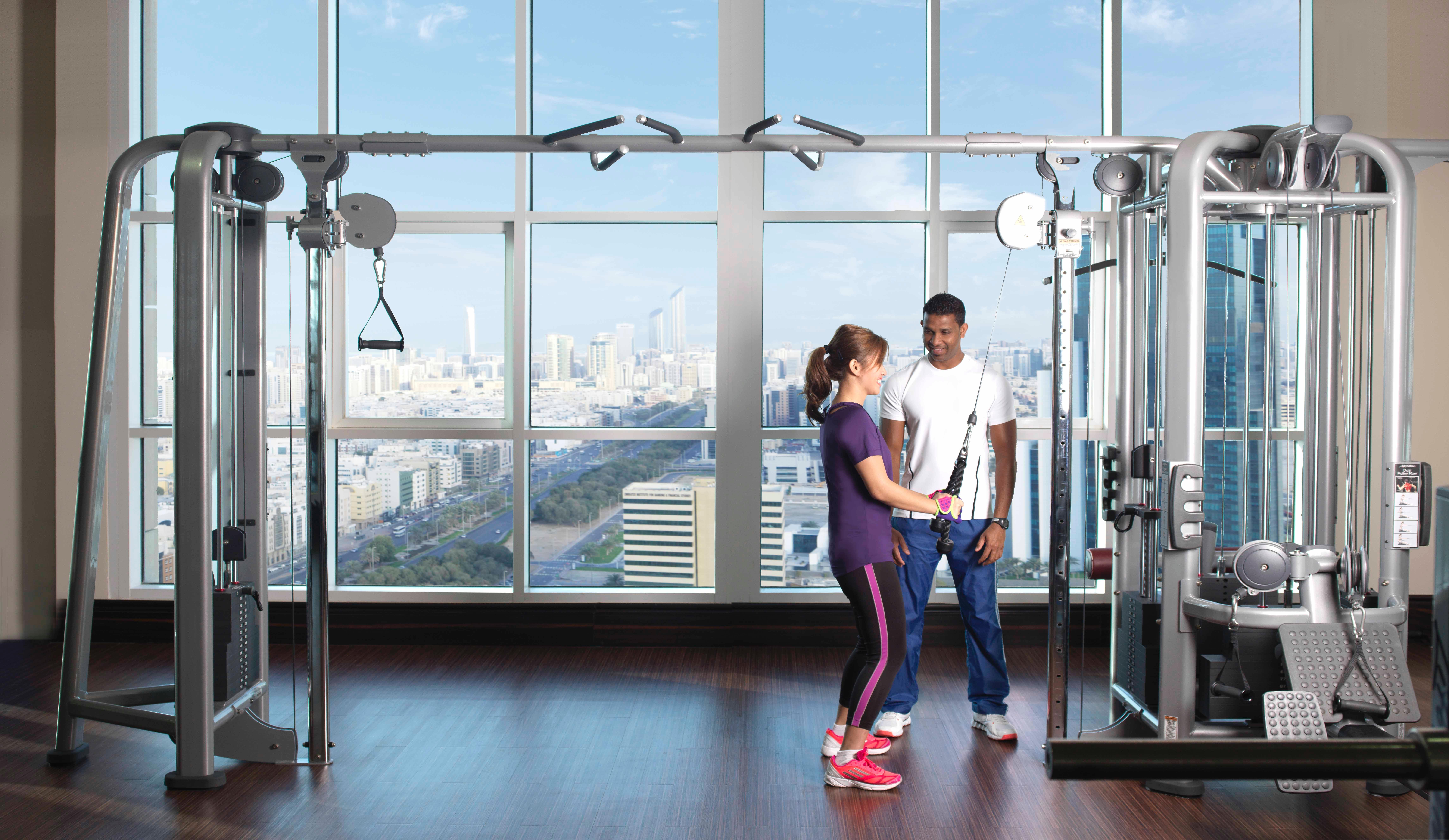 Dusit Thani Abu Dhabi Exterior photo The photo shows a modern gym setting with large windows offering a view of a cityscape. In the foreground, there are two individuals engaged in a training session. One person, a woman in athletic wear, appears to be exercising with a machine, while a