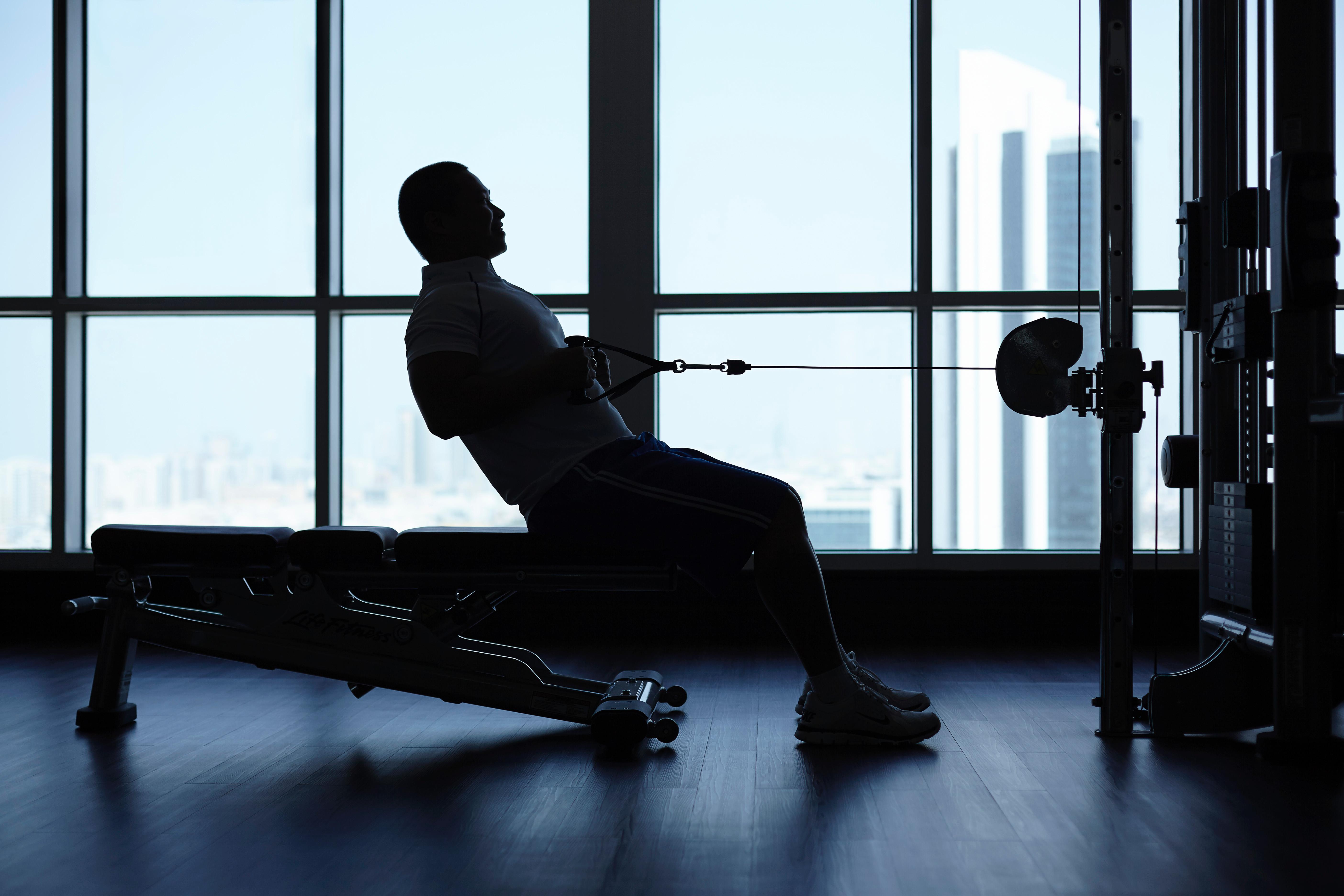Dusit Thani Abu Dhabi Exterior photo The photo shows a silhouette of a person exercising in a gym. The individual is seated on a bench, using a cable machine, and appears to be performing a pulling exercise. The backdrop features large windows that provide a view of a cityscape, with ta