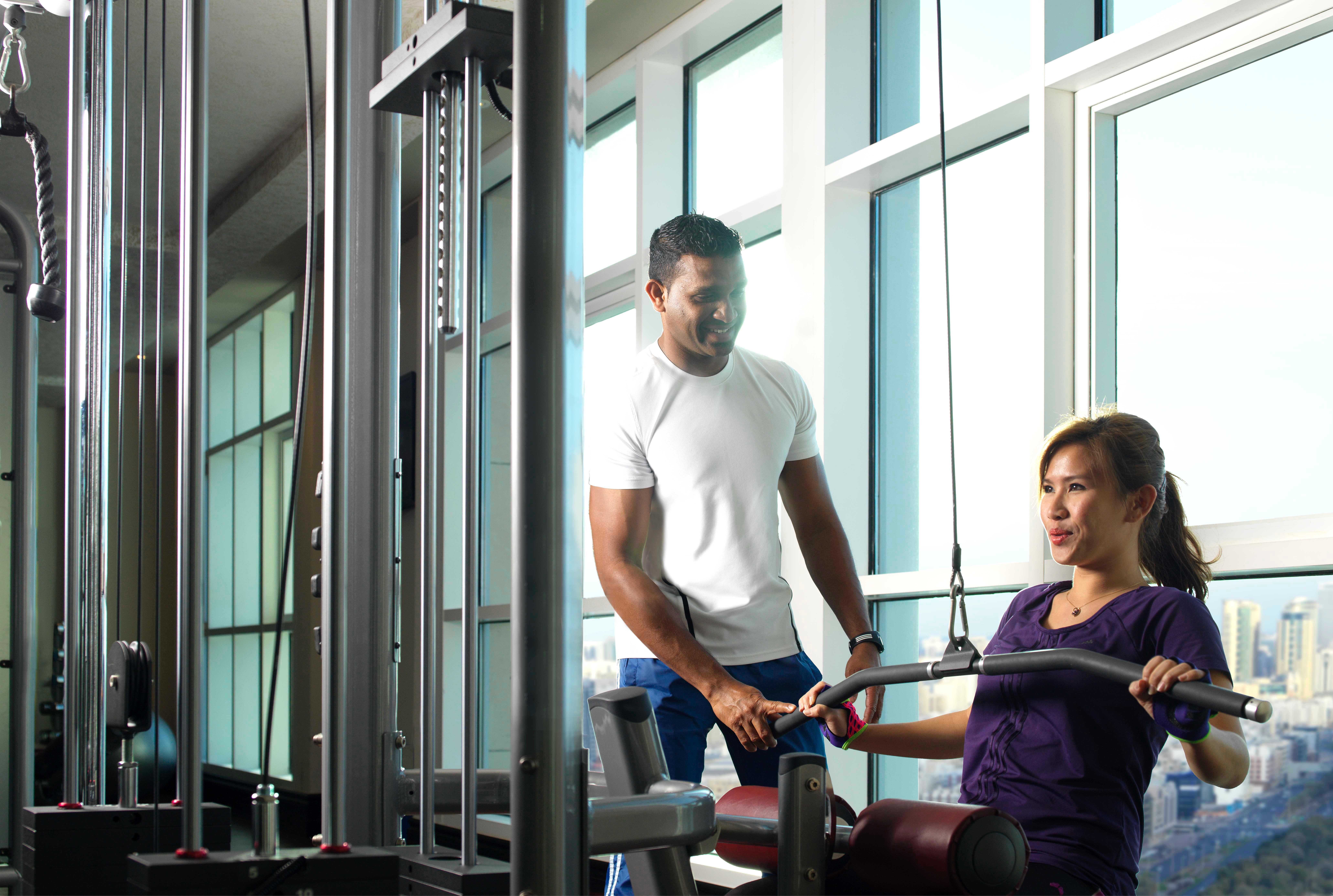 Dusit Thani Abu Dhabi Exterior photo The photo shows two individuals in a gym setting. One person, likely a trainer, is assisting a woman who appears to be exercising on a machine. The gym has large windows that let in natural light, creating a bright atmosphere. The trainer is focused 