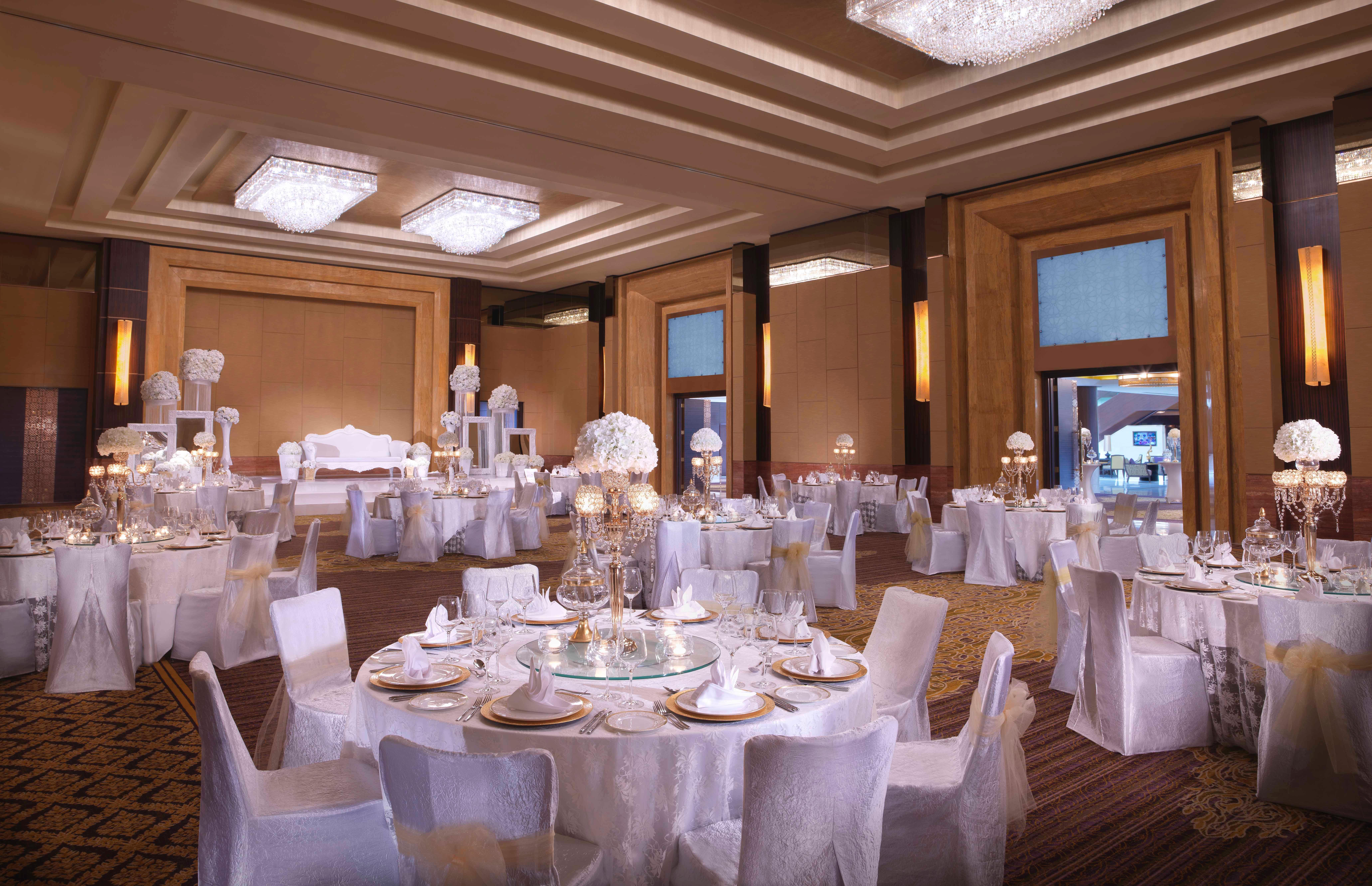 Dusit Thani Abu Dhabi Exterior photo The photo shows an elegantly decorated banquet hall arranged for an event, likely a wedding or formal celebration. The room features round tables covered with white tablecloths, each set with fine dining ware, including plates and glasses. The chairs