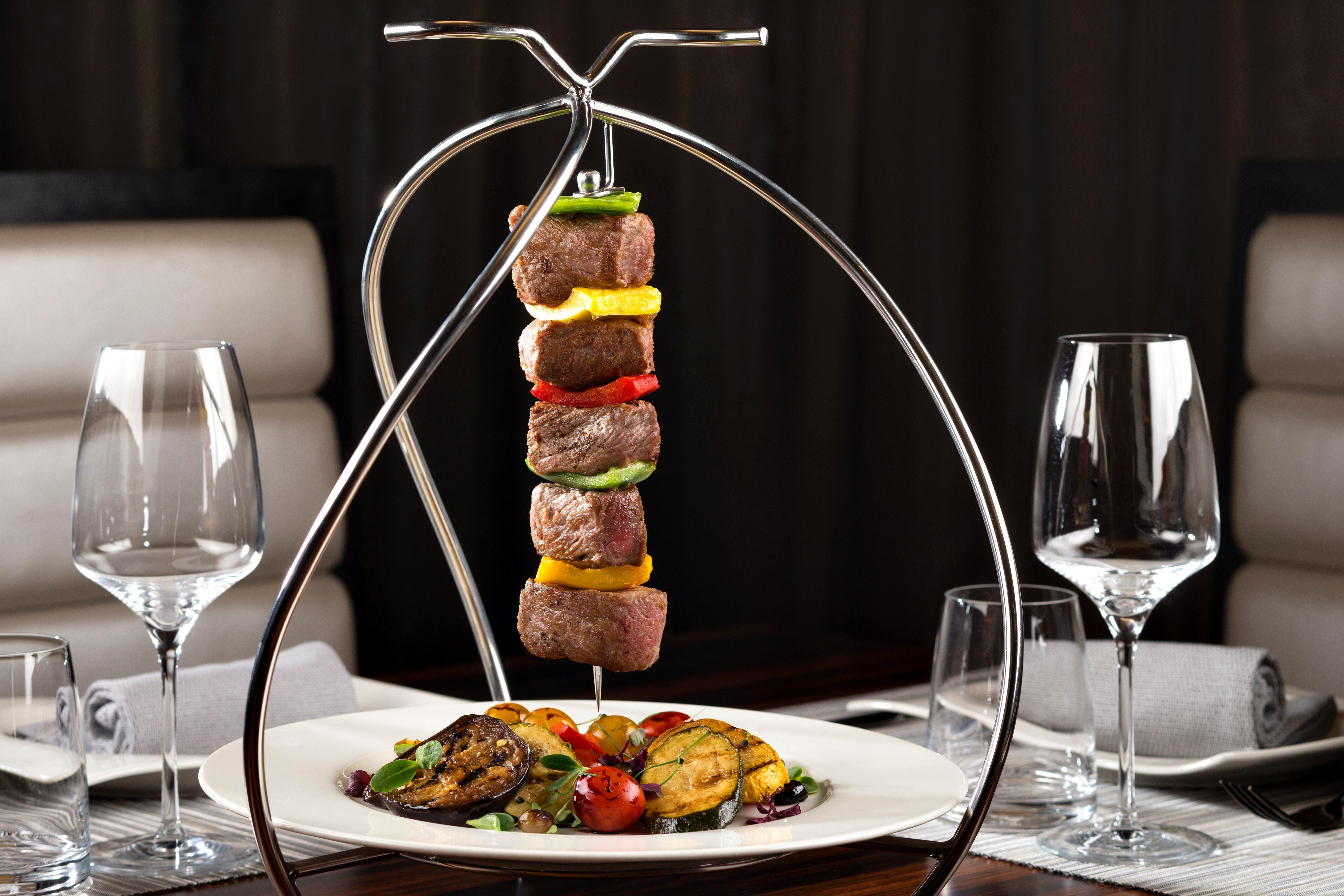 Dusit Thani Abu Dhabi Exterior photo The photo shows a stylish presentation of skewered meat and vegetables. The skewers are hanging from a metallic stand, featuring chunks of beef interspersed with colorful bell peppers and onions. Below the skewers, there is a plate that includes gril
