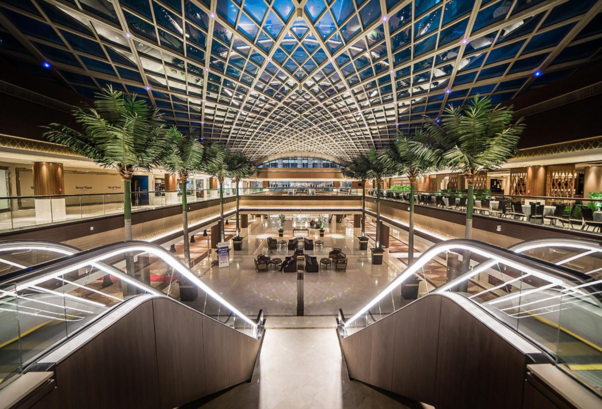 Dusit Thani Abu Dhabi Exterior photo The photo shows a large, modern interior space with an impressive glass ceiling featuring a geometric design. The area is illuminated with soft lighting, highlighting various levels and open spaces. Tall indoor palms add a touch of greenery to the en