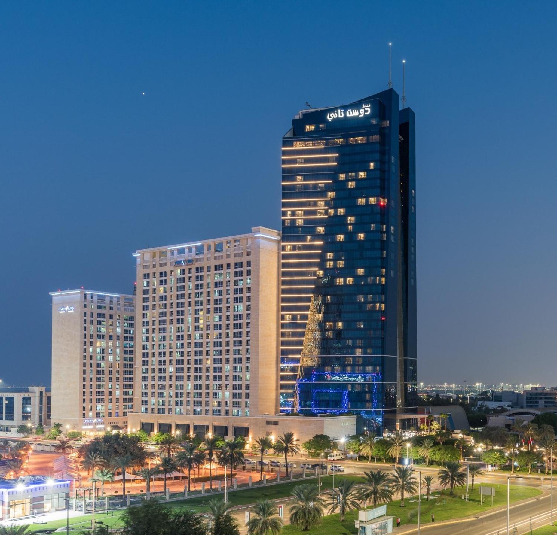 Dusit Thani Abu Dhabi Exterior photo The image shows a tall modern hotel building illuminated at night. There are two distinct sections: a low-rise building on the left and a high-rise tower on the right. The hotel is adorned with bright lights that highlight its architectural features,