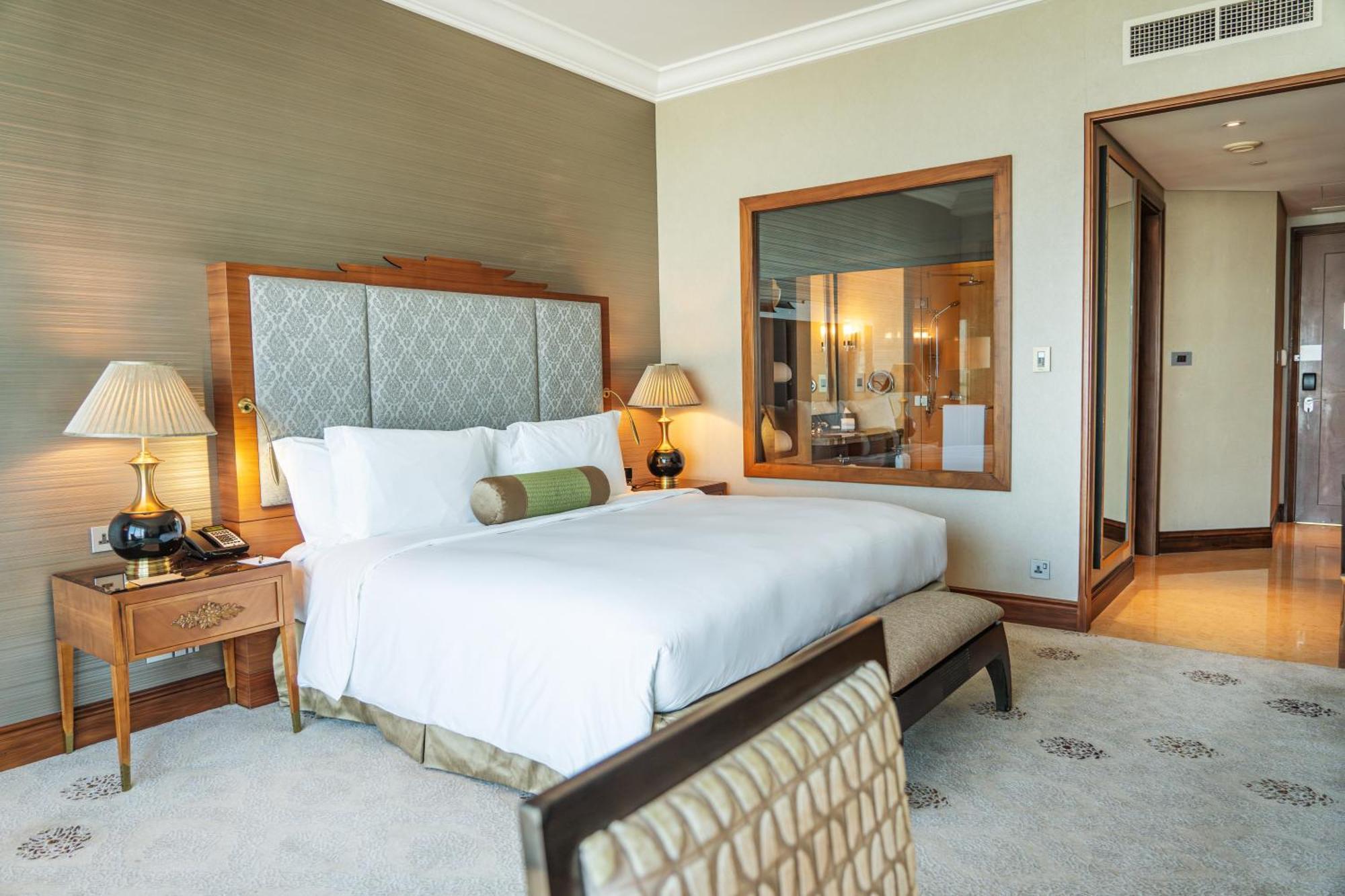 Dusit Thani Abu Dhabi Exterior photo The photo depicts a well-appointed hotel room featuring a large bed with white linens and decorative pillows. On either side of the bed, there are stylish bedside tables with lamps. The room has a warm color palette, accentuated by a textured wall. I