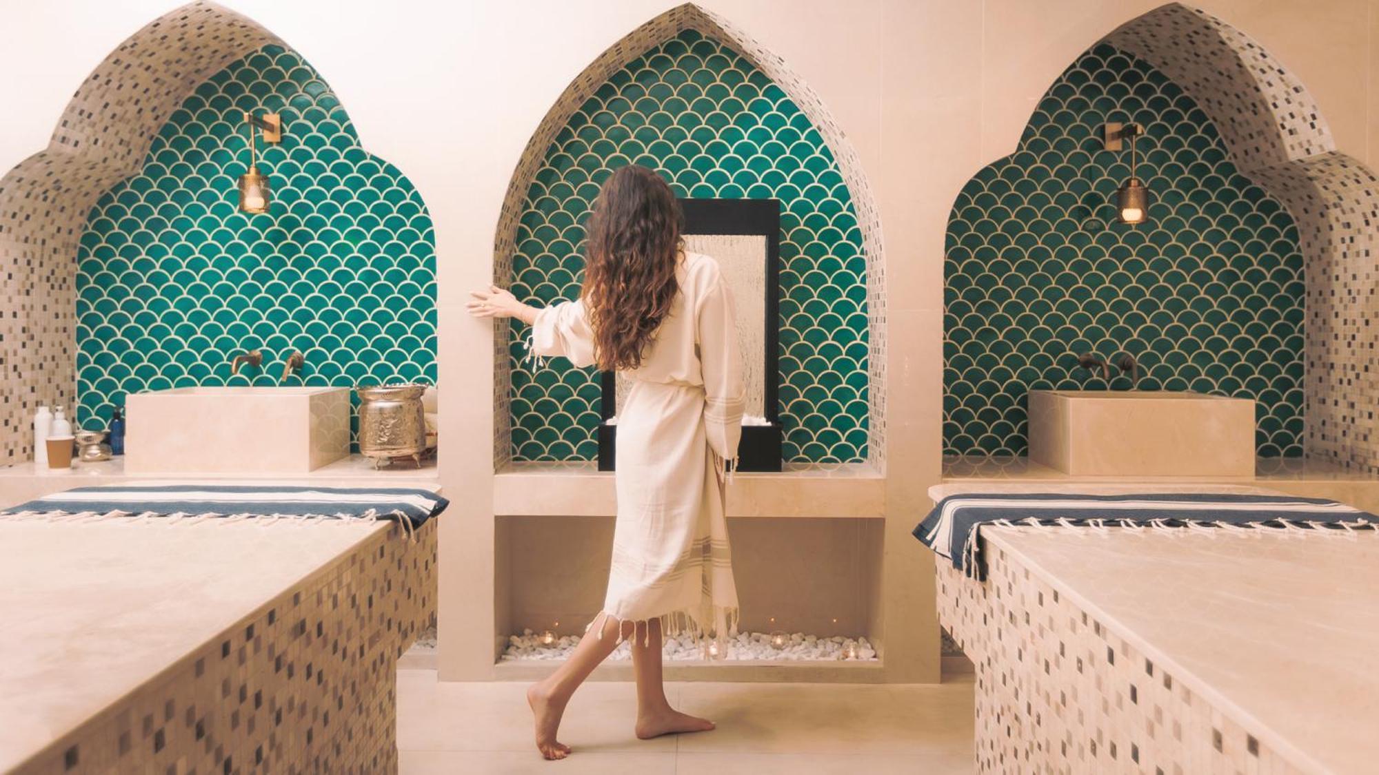 Dusit Thani Abu Dhabi Exterior photo The image shows a serene spa setting with a person standing in the foreground, dressed in a light robe. The background features elegant arched niches adorned with intricate teal tile work, giving a Moroccan-inspired aesthetic. The space is decorated 